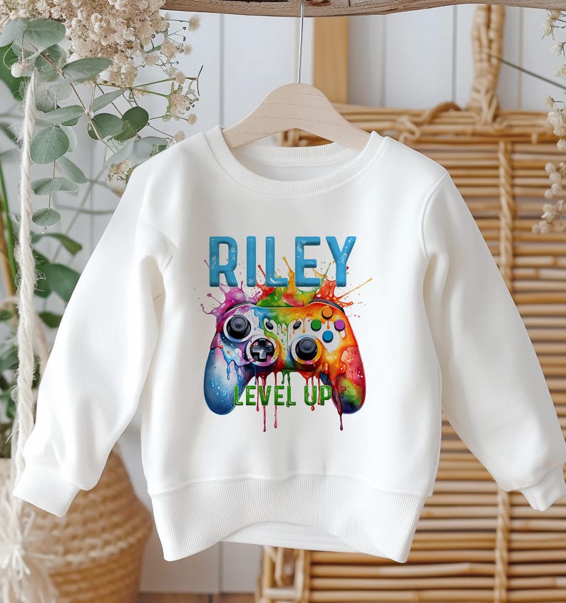 K&ouml;nnte beinhalten: Wei&szlig;es Sweatshirt mit einer farbenfrohen Videospiel-Controller-Grafik und dem Namen "RILEY" dar&uuml;ber. Der Controller ist eine lebendige Mischung aus Farben, mit den Worten "LEVEL UP" darunter. Das Design hat einen Splash-Art-Stil.