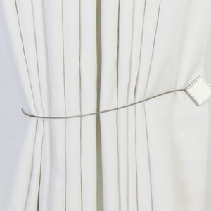 May include: White curtains tied back with a silver metal tieback. The tieback has a square shape with rounded corners.