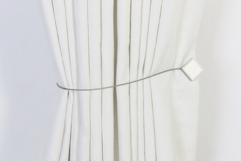 May include: White curtains tied back with a silver metal tieback. The tieback has a square shape with rounded corners.