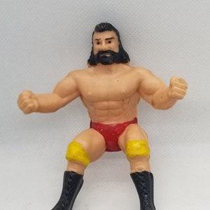 May include: A plastic action figure of a wrestler wearing a red and yellow wrestling outfit. The figure has a black beard and black hair.