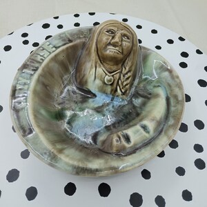May include: A ceramic ashtray with a Native American figure in relief. The ashtray is decorated with brown, green, and blue glaze. The figure has a feathered headdress and a beaded necklace.