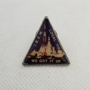 May include: A triangular metal pin with a blue and white image of a rocket launch. The text "I.B.E.W. LOCAL 110 WE GOT IT UP" is printed on the pin.