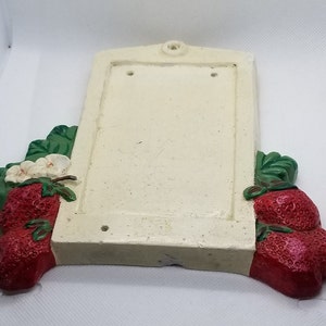 May include: A white ceramic sign with a blank rectangular space in the center. The sign is decorated with green leaves and red strawberries on the sides.