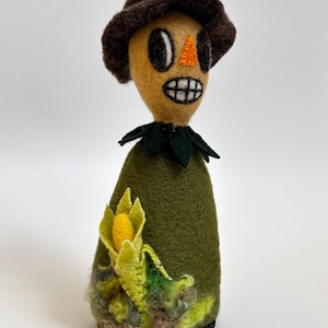 May include: A needle-felted scarecrow figurine with a dark brown hat, a tan face, an orange triangular nose, and a wide toothy grin. The body is olive green with a dark green collar and a textured yellow corn cob accent at the base.