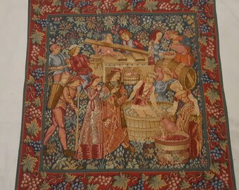 May include: A tapestry depicting a group of people working in a vineyard. The tapestry is framed with a red and green border with grapes and leaves. The people are dressed in medieval clothing and are harvesting grapes, pressing grapes, and carrying baskets of grapes.