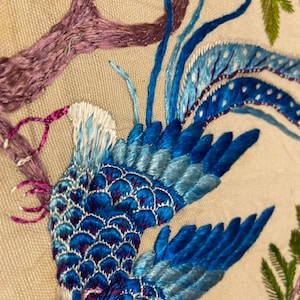 May include: An embroidered bird, blue and white in colour, with a long tail, perched on a branch with pink flowers. The bird is surrounded by green leaves and branches.