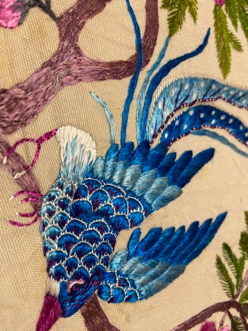 May include: An embroidered bird, blue and white in colour, with a long tail, perched on a branch with pink flowers. The bird is surrounded by green leaves and branches.