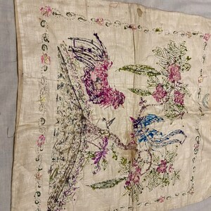 May include: A white fabric square with a hand-stitched design of two birds and a flower. The birds are blue and purple, and the flower is pink and green. The fabric is surrounded by a border of pink flowers.