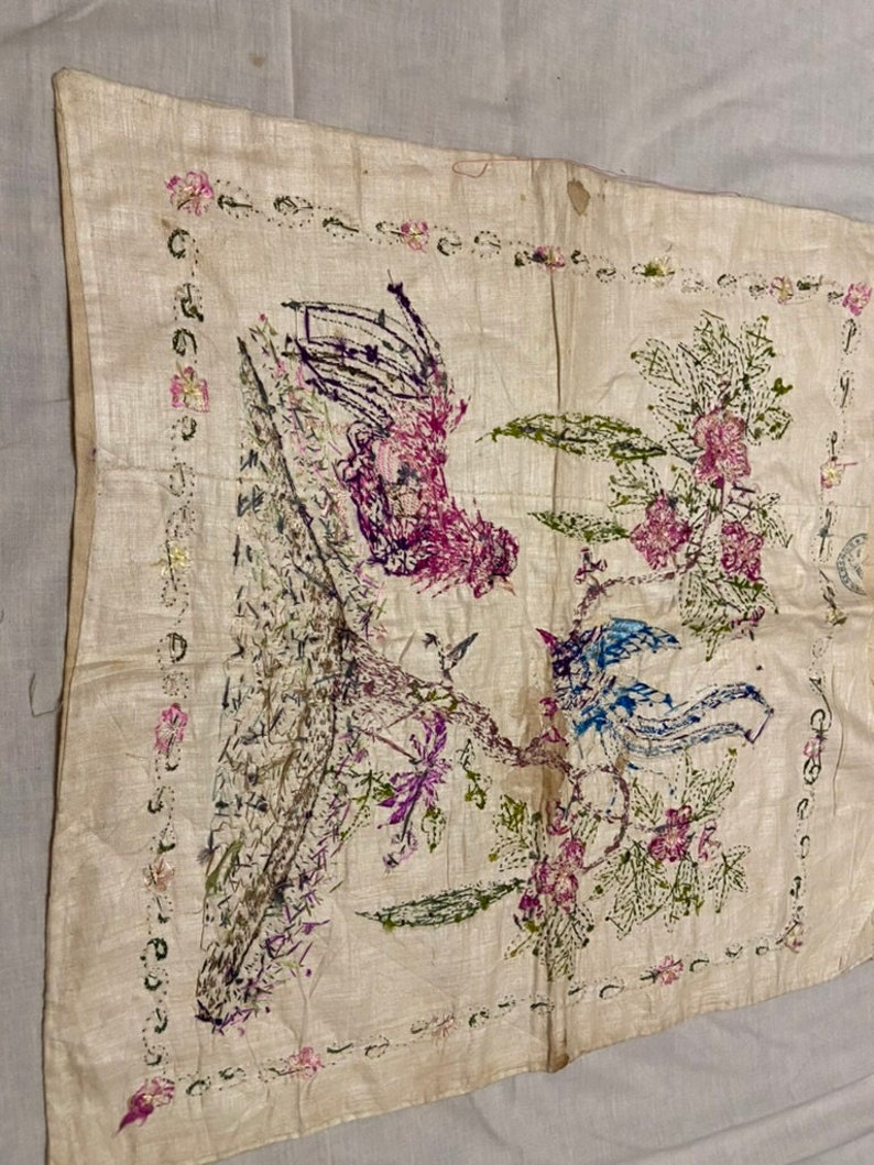 May include: A white fabric square with a hand-stitched design of two birds and a flower. The birds are blue and purple, and the flower is pink and green. The fabric is surrounded by a border of pink flowers.
