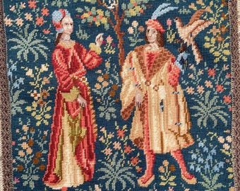May include: A needlepoint tapestry depicting a man and woman standing in a garden with a tree and birds. The tapestry is framed with a brown and gold trim.