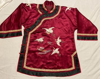 May include: A burgundy satin jacket with black trim and gold accents. The jacket features embroidered white cranes and cloud designs. The sleeves are three-quarter length, and the jacket has a mandarin collar.