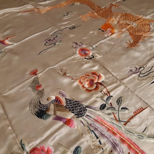 May include: A cream-coloured silk fabric with intricate embroidery of a phoenix, dragon, and floral designs. The phoenix is depicted in shades of brown, black, and purple, while the dragon is orange and yellow. The floral designs are in shades of pink and red.