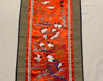 May include: A red silk tapestry with a border of brown fabric. The tapestry features an embroidered scene of white cranes flying over a red and green landscape. The Chinese characters "大吉" are embroidered in black thread in the upper right corner of the tapestry.