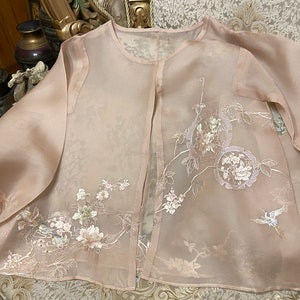 Può includere: Giacca trasparente rosa pallido con maniche lunghe e parte anteriore aperta. È adornata con ricami floreali e di uccelli in bianco, rosa e verde. Il tessuto delicato e i dettagli intricati suggeriscono un capo formale o per occasioni speciali.