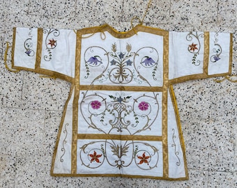 May include: A white garment with gold trim and intricate floral embroidery in shades of pink, purple, and yellow. The garment has a long, flowing design and is likely a religious robe.