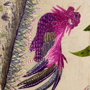 May include: Embroidered design of a pink bird with a long tail, with green and purple accents, on a beige fabric background.