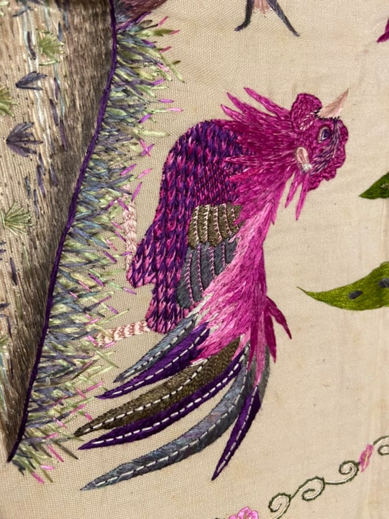 May include: Embroidered design of a pink bird with a long tail, with green and purple accents, on a beige fabric background.