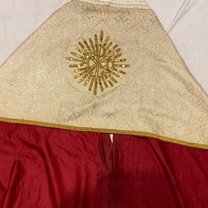 May include: A white and gold liturgical vestment with a red lining. The vestment features a gold embroidered design with a cross in the center.