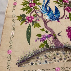 May include: A beige fabric square with an embroidered design of two birds perched on branches with pink flowers and green leaves. The border is a green and white floral design.