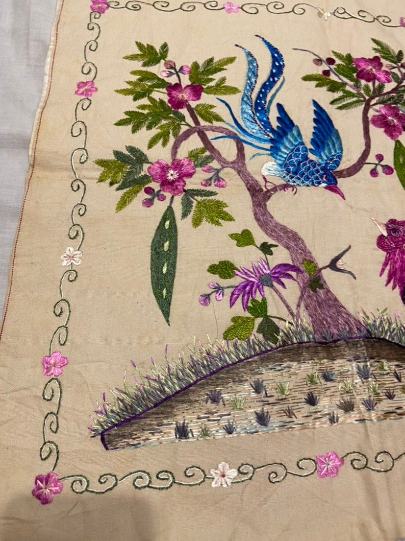 May include: A beige fabric square with an embroidered design of two birds perched on branches with pink flowers and green leaves. The border is a green and white floral design.