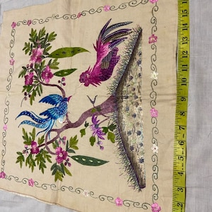 May include: A beige fabric square with embroidered birds and flowers. The birds are a blue bird with a long tail and a pink rooster. The flowers are pink and white.