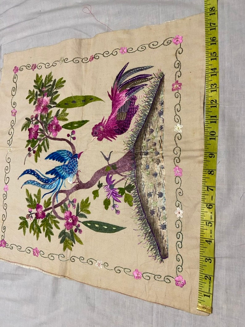 May include: A beige fabric square with embroidered birds and flowers. The birds are a blue bird with a long tail and a pink rooster. The flowers are pink and white.