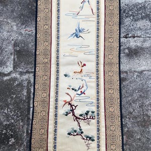 May include: Embroidered wall hanging depicting a tranquil landscape with a mountain, birds, and a tree branch. The artwork is framed with a decorative border in gold, blue, and red. The background is cream-colored.
