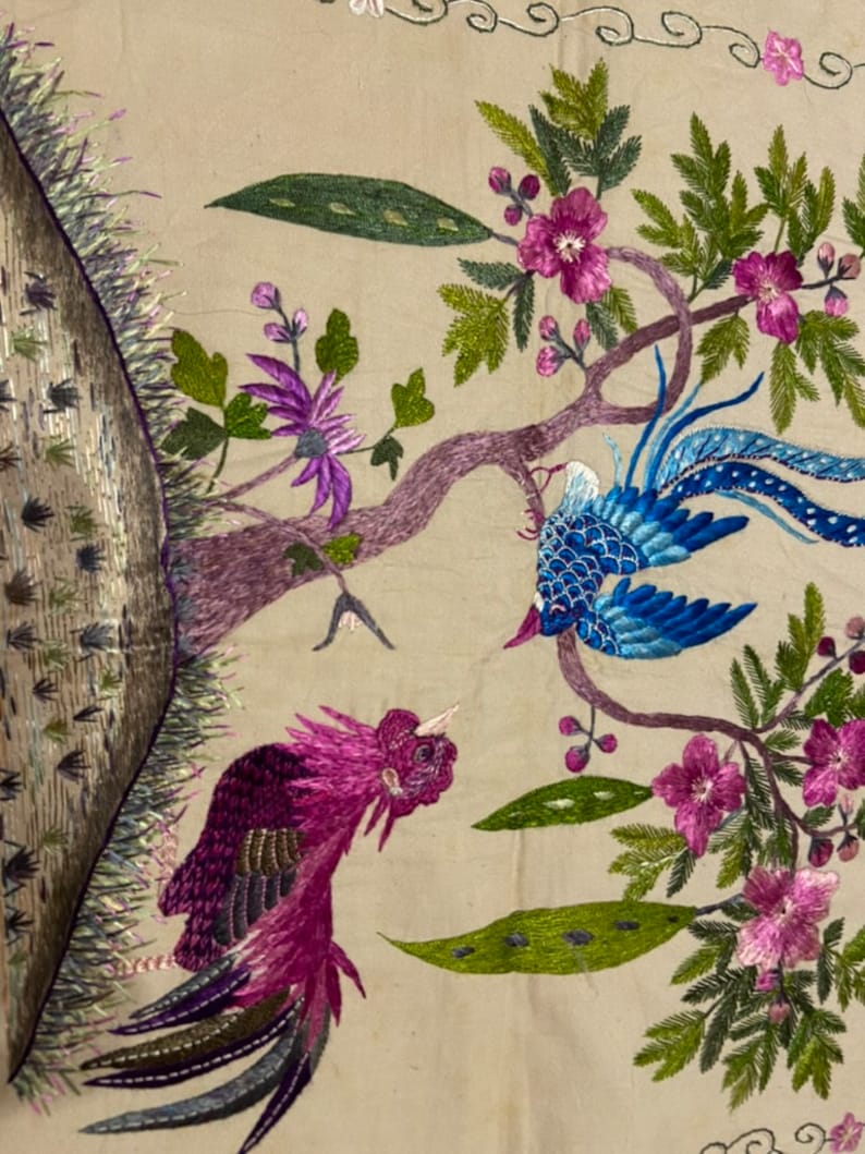 May include: Embroidered wall hanging featuring two birds with colourful plumage, perched on a branch with pink flowers and green leaves. The background is a beige fabric.
