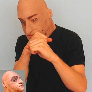 Può includere: Una maschera per la testa calva dall'aspetto realistico con rughe dettagliate e la fronte corrugata. La maschera è realizzata in un materiale liscio color carne e ha un aspetto realistico.