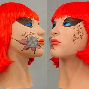 May include: A mannequin head with bright red hair styled in a bob. The face is painted with a blue flower and black and brown vines on the cheek. The lips are painted a bright orange.