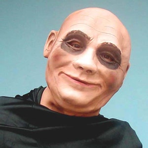 May include: A close-up of a bald man wearing a black mask with dark makeup around the eyes. The mask has a painted-on smile and appears to be made of latex.