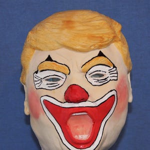 Könnte beinhalten: Eine Clownmaske mit einem blassen Gesicht, einer roten Nase und einem breiten roten Mund, der schwarz und weiß umrandet ist. Die Maske hat blondes Haar und bemalte Augenbrauen und Augendetails. Die Wangen sind rosa bemalt, und die Maske ist vor einem blauen Hintergrund platziert.