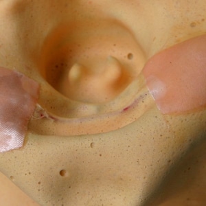 May include: Close-up of a light brown textured surface with two light pink adhesive bandages. The bandages are rectangular and have a slightly textured surface.