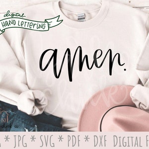 May include: A white sweatshirt with the word "amen" written in black cursive script. The sweatshirt is folded and laid on a wooden surface with a pair of blue jeans and a pink hat.