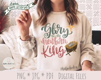 Puede incluir: Una sudadera blanca con un diseño navideño. El diseño presenta las palabras "Gloria al recién nacido Rey" en letras verdes, rosas y amarillas. También se representa un pesebre con un bebé Jesús dentro del diseño.