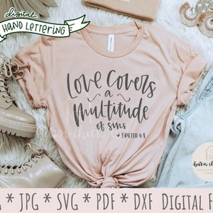 Op de afbeelding: Een lichtroze t-shirt met de handgeschreven tekst "Love covers a multitude of sins". De afbeelding bevat een paar lichtgekleurde laarzen en een jeans. De woorden "digital hand lettering" zijn ook zichtbaar.