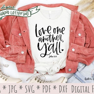 May include: A white t-shirt with the text "Love one another, Y'all" in black script lettering. The text "John 15:39" is also included in black lettering. The t-shirt is laid out on a white surface with a red corduroy jacket, a pair of white boots, and a pair of blue jeans.