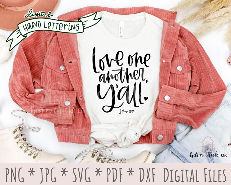 May include: A white t-shirt with the text "Love one another, Y'all" in black script lettering. The text "John 15:39" is also included in black lettering. The t-shirt is laid out on a white surface with a red corduroy jacket, a pair of white boots, and a pair of blue jeans.