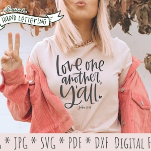 May include: A white t-shirt with the text "Love one another, Y'all" in black script font. The text "John 13:34" is also included in smaller font. The shirt is being worn by a person with a pink jacket.