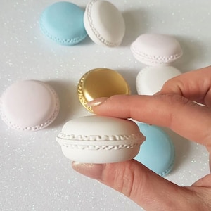 Può includere: Assortimento di sei piccoli macarons rotondi di colore pastello. I macarons sono disposti su una superficie bianca e scintillante. Un macaron è tenuto da una mano.