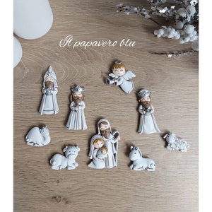 May include: A white nativity scene set with figurines of Mary, Joseph, baby Jesus, an angel, three wise men, a camel, a sheep, and a donkey. The figurines are all white with painted details and are arranged on a wooden surface. The text "Il papavero blu" is written in the upper center of the image.