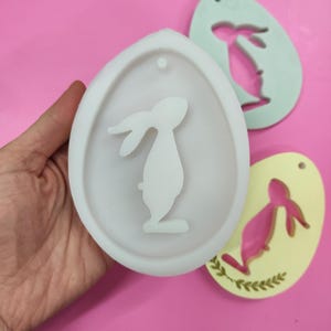 May include: White silicone mold shaped like an egg with a bunny silhouette inside. The mold has a small hole at the top for hanging.