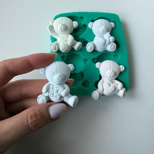 Può includere: Una mano tiene in mano una piccola statuina di orsetto bianco. L'orsetto è una delle quattro figure simili in uno stampo in silicone verde acqua. Gli altri orsetti sono anch'essi bianchi e in varie pose.