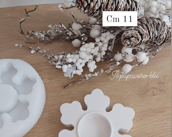 May include: White snowflake-shaped candle holder made of plaster. The holder is 11 cm in diameter and has a circular opening in the center for a candle. The holder is sitting on a wooden surface.