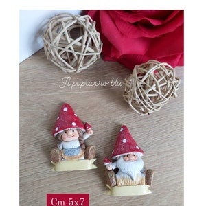 Puede incluir: Dos pequeñas figuras de gnomos con sombreros de hongo rojo y blanco. Los gnomos están sentados en una pancarta amarilla y sostienen un hongo. Los gnomos miden 5 cm por 7 cm.