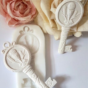 Puede incluir: Dos moldes de silicona blancos en forma de llave con un cáliz y un diseño de trigo. La llave tiene las letras "JHS" en ella.