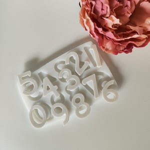 Può includere: Stampo in silicone bianco con i numeri da 0 a 9 e da 1 a 7. Lo stampo è su una superficie bianca.