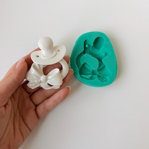 May include: A white pacifier-shaped object featuring a decorative bow at the base and a matching teal silicone mould. The pacifier has a round nipple, a shield with two small holes, and a sculpted bow detail attached to the ring portion.