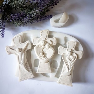 May include: A white silicone mold featuring three crosses with religious figures. The crosses depict Jesus, the Holy Family, and an angel. The mold is on a white surface with purple floral accents.