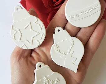May include: Four white ceramic Christmas ornament shapes, each with a different design. The ornaments are 6 cm by 4 cm and are held in a hand. The designs include a star, a reindeer, a snowflake, and the words "Merry Christmas!"